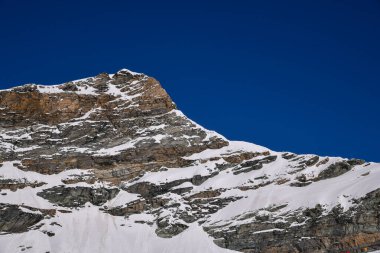 Derin mavi gökyüzüne karşı engebeli, karlı bir dağın dramatik yakın görüntüsü. Keskin kaya dokuları, kış manzarası ve alp atmosferi seyahat, doğa ve macera temaları için çarpıcı bir doğal manzara yaratır..