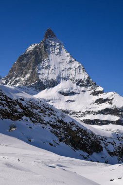 Matterhorn 'un görkemli manzarası, derin mavi gökyüzünün altında karlı dağlık yamaçların üzerinde yükseliyor. Dramatik kış ışığı engebeli kaya yüzeyini ve el değmemiş dağ manzarasını vurguluyor..