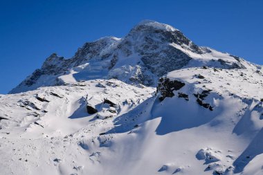Derin mavi bir gökyüzünün altında engebeli dağlık yamaçlardan yükselen dramatik karlı dağ zirvesi. Parlak güneş ışığıyla kış manzarası, kayalık detaylar, ve el değmemiş kar sakin, görkemli bir atmosfer yaratır..
