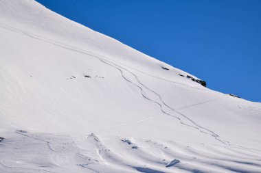 Derin mavi bir gökyüzünün altında, karlı bir yamaçta taze kayak izleri kıvrılıyor. Tepeden çekilen kış manzarası, el değmemiş toz ve sakin alp manzarasını gösteriyor..