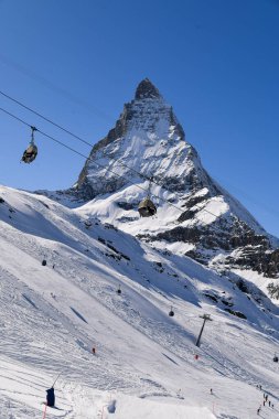 İsviçre 'nin Zermatt kentindeki ikonik Matterhorn zirvesinin dikey kış manzarası. Derin mavi bir gökyüzünün altında, kar yamaçlarında hareket eden modern kayak kaldırma koltukları. Kış sporları ve seyahat temaları için manzara dağları.