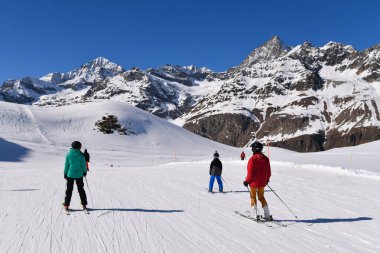 Kayakçılar, arka planda muhteşem İsviçre dağ zirveleri bulunan manzaralı bir dağ yamacında güneşli bir kış gününün tadını çıkarıyorlar. Açık mavi gökyüzü, taze kar ve nefes kesici manzaralar mükemmel bir kayak atmosferi yaratır..