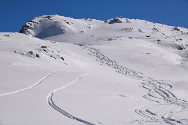 Derin mavi bir gökyüzünün altında, karlı bir yamaçta taze kayak izleri kıvrılıyor. Tepeden çekilen kış manzarası, el değmemiş toz ve sakin alp manzarasını gösteriyor..