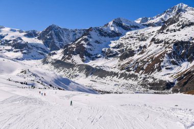 İsviçre Alpleri 'ndeki geniş karlı vadi yamaçtan geçen kayakçılarla dolu. Görkemli dağ zirveleri, derin mavi bir kış gökyüzünün altında yükselerek, dağlık doğanın güzelliğini yakalar..