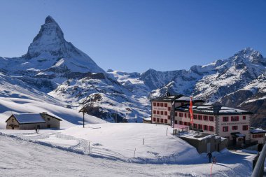 Matterhorn 'un manzarası ve Zermatt, Swtthe' de bir dağ oteli. Parlak mavi gökyüzü, karlı yamaçlar, dağlık binalar ve kayakçılar kışın İsviçre Alpleri 'nin huzurlu bir atmosferini oluşturur..
