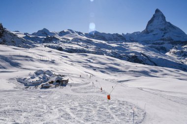Karlı dağ manzarası, panoramik kış yamaçları, uzakta kayakçılar, ve berrak mavi gökyüzünün altında dağların üzerinde yükselen ikonik Matterhorn zirvesi. Huzurlu İsviçre kış manzarası.