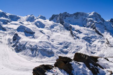 Muazzam karlı bir buzulu, engebeli zirveleri ve berrak mavi gökyüzünün altında derin yarıkları olan dramatik alp manzarası. Kış dağ manzarası ham doğa, buzlu dokular ve yüksek irtifa arazilerini yakalıyor..