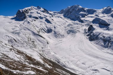 Alplerde parlak mavi gökyüzü, dik yamaçlar ve buzul oluşumlarıyla görkemli karlı dağ sırtı. Çarpıcı bir kış manzarası yüksek irtifa doğasının ham güzelliğini yansıtıyor..