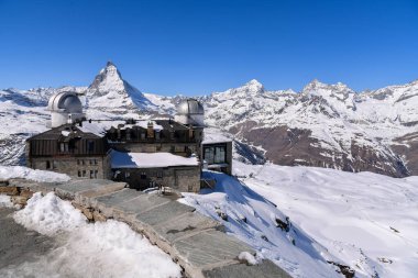 Arkasında ikonik Matterhorn zirvesi olan karla kaplı Gornergrat Gözlemevi. Açık mavi gökyüzü, dramatik alp manzarası, kış dağları ve İsviçre Alplerinde el değmemiş kar..