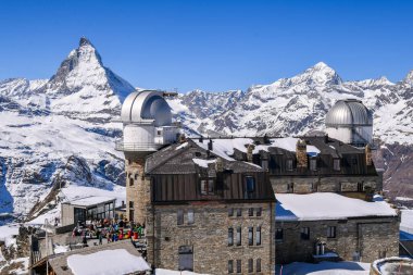 Arkasında ikonik Matterhorn zirvesi olan karla kaplı Gornergrat Gözlemevi. Açık mavi gökyüzü, dramatik alp manzarası, kış dağları ve İsviçre Alplerinde el değmemiş kar..