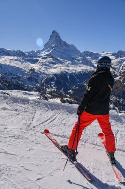 Kırmızı pantolonlu bir kayakçı, parlak bir kış gününde, ikonik madde boynuzu ve İsviçre Alpleri 'nin çevresini net bir şekilde gören karlı bir yamaçta duruyor..