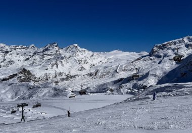 İsviçre Alpleri 'ndeki karlı bir dağ yamacından inen kayakçılar. Koltuk asansörleri ve arka planda dramatik kış zirveleri. Aktif kış sporları ve dağlık arazi.