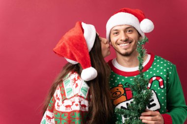 Çirkin bir kazak giyen ve elinde küçük bir Noel ağacı tutan İspanyol bir adam kız arkadaşının yanağından öpüyor.