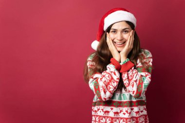 Noel Baba şapkalı, çirkin kazaklı, gülümseyen ve yüzüne dokunan güzel bir kadın kırmızı arka planlı bir stüdyoda.