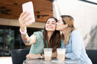 Lezbiyen beyaz kadınlar, selfie çekerken İspanyol genç kız arkadaşının yanağını öpüyor. 