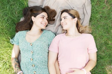 İki mutlu kız arkadaş parkta battaniyenin üzerinde uzanırken göz teması kurup el ele tutuşuyorlar.