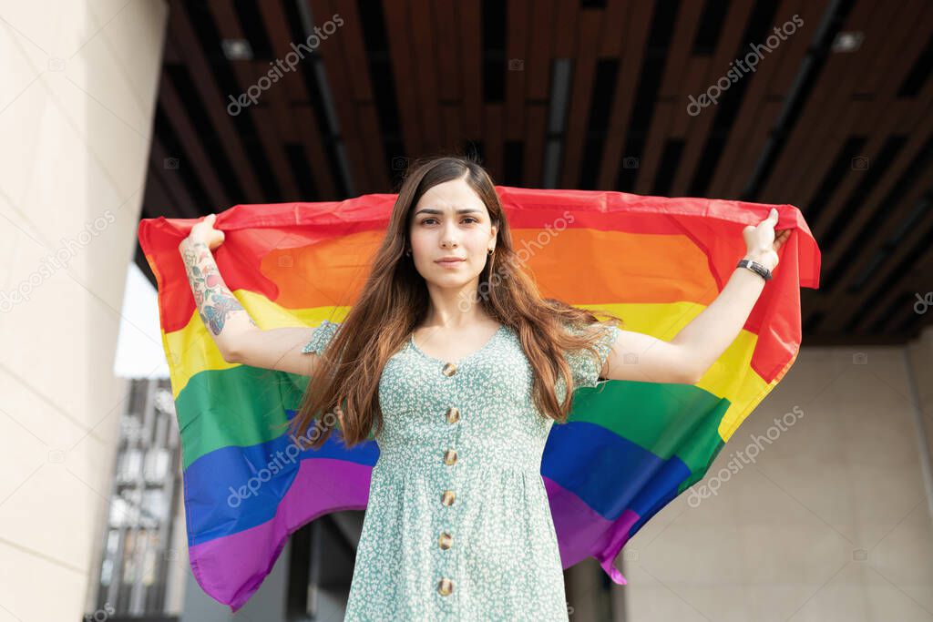 Portrait of an attractive hispanic woman holding a LGBT rainbow flag behind her back and looking serious