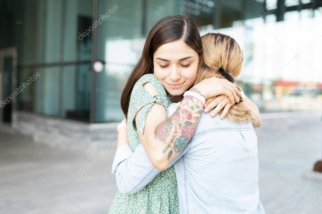 Latin young gay woman looking peaceful and happy while embracing her blond lesbian girlfiend outside a building