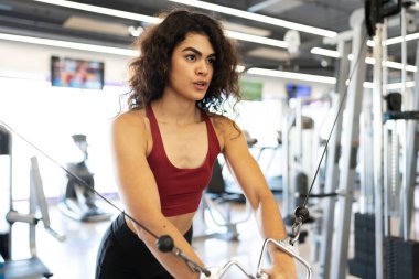 Spor salonunda kollarını ve göğüs kaslarını kablo makinesiyle çalıştıran genç bir kadının yan görüntüsü.