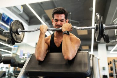 Spor salonunda halterle ağırlık kaldırırken kol kaslarını ve pazılarını çırpan atletik genç bir adam.