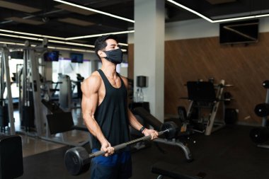 Yüz maskeli yakışıklı genç adam spor salonunda halterle ağırlık kaldırma egzersizi yapıyor.