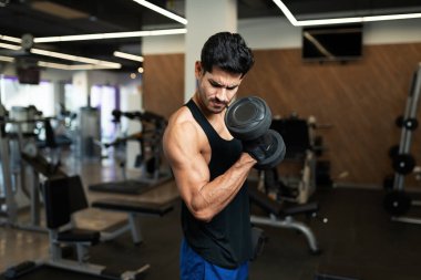 Çekici Latin genç adam kaslarını esnetiyor ve spor salonunda ağırlık kaldırıyor.