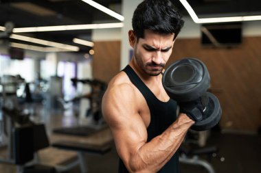 İspanyol genç adam vücut geliştirme merkezinde ağırlık kaldırırken kol kaslarına bakıyor.
