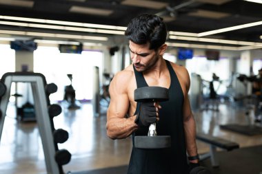 Modern bir spor salonunda ağırlık kaldırırken bisepslerine bakan yakışıklı Latin bir genç.