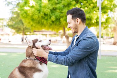 Yakışıklı bir Latin yetişkinin profili mutlu görünüyor ve yeşil çimlerde köpeğiyle göz teması kuruyor. 
