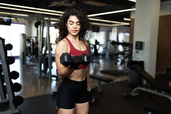 运动的拉丁裔年轻女子在健身房里做二头肌卷和举重时 看着她的手臂肌肉 — 图库照片
