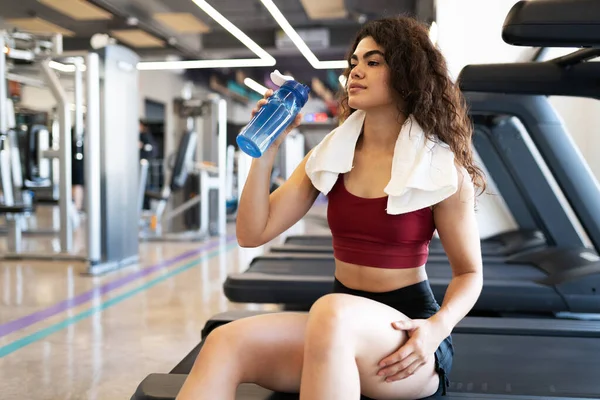 Latin woman in her 20s drinking water and wearing a towel after running in a treadmill at the fitness center