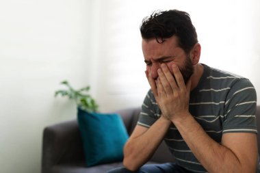 Latin yetişkin bir adam, üzgün ve üzgün bir şekilde kanepede oturup ağlarken elleriyle hasılatı örtüyor.