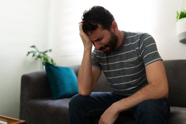 Hispanic man holding his head with one hand while sitting on the sofa and crying, looking sad and depressed