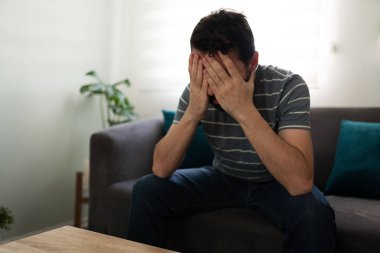 Hispanic man covering his face with his hands, looking sad and depressed on the couch