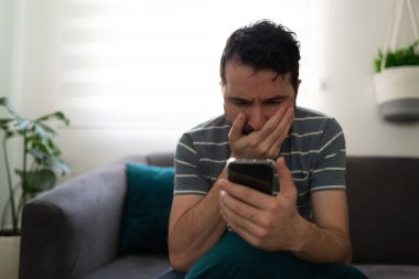 Latin bir adam cep telefonuna bakıp koltukta tek başına otururken şaşırmış görünüyor.