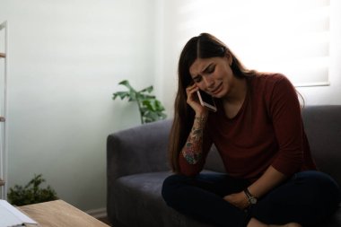 Attractive hispanic young woman crying and talking on the phone about her troubles. Depressed and lonely woman in the living room clipart