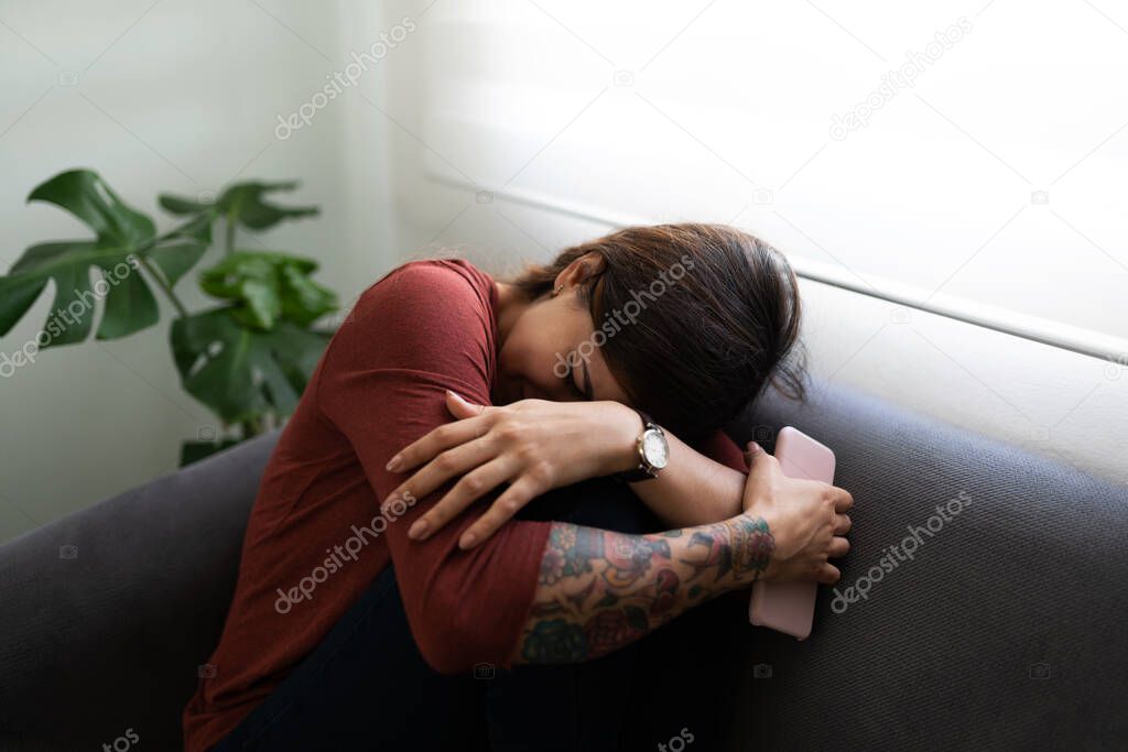 Sad young woman crying and hugging her knees on the couch. Latin woman suffering because of a break up 