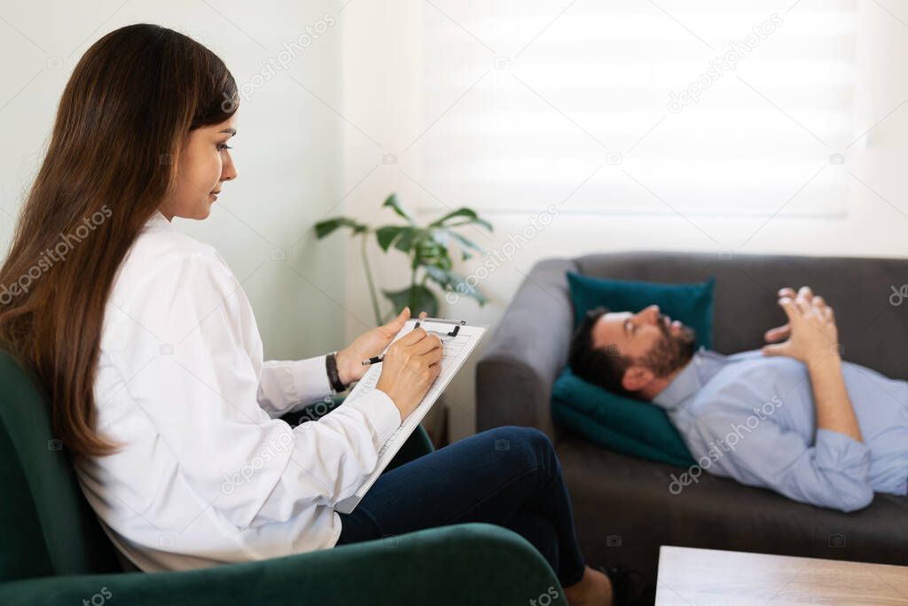 Young female pshycologist writing on her chart during a therapy session with her male patient 