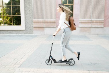 30 'lu yaşlarında bir kadının binanın dışında scooter sürerken görüntüsü. Kafkasyalı kadın işe gitmek için yeşil bir araç kullanıyor.
