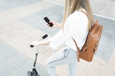 Sırt çantalı bir kadın çalışanın akıllı telefonunu kullanırken ve scooterını tutarken çekilmiş fotoğrafı, çevre dostu bir araç. 