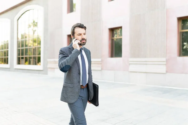 ラテン語の男のフロントビューを歩いて 彼の電話で話しています 都市の建物の外にポートフォリオを持っているプロの男 — ストック写真