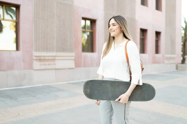 スケートボードを運ぶ街の建物に到着魅力的な女性 代替輸送手段を選択する女性従業員 — ストック写真