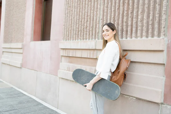 一个漂亮的女人拿着滑板站在一座城市的建筑外的画像 使用环保车辆的背包的女雇员 — 图库照片