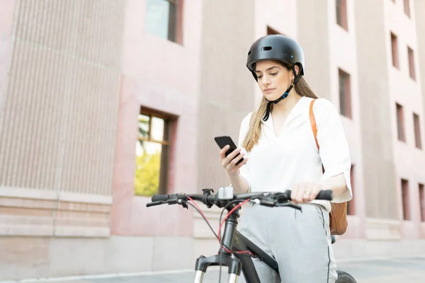 オフィスビルの外でスマホにテキストメッセージを送る格好いい女性 エコカーとして自転車用ヘルメットを使用する女性労働者 — ストック写真