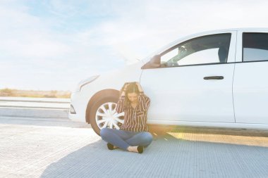 Elleri kafasında, yolda bozuk arabasının yanında oturan endişeli genç bir kadın. Genç bir kadın otoyolda çekici bekliyor. 