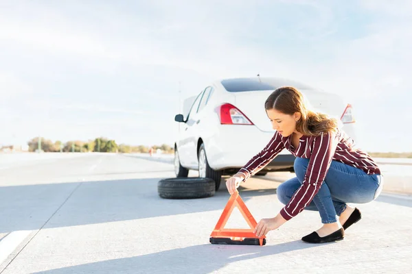 彼女の車は平らなタイヤで道端に駐車されているので 若い女性は警告として通りに反射三角形の標識を置きます — ストック写真