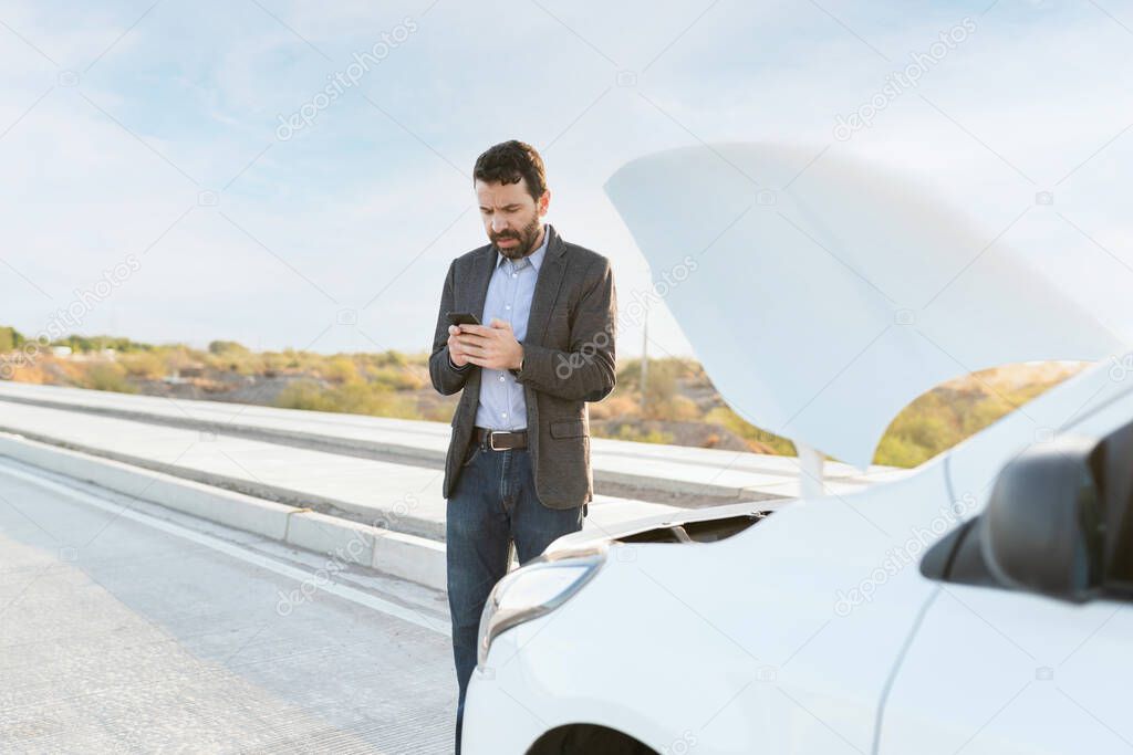 Latin man in his 30s with a dead car texting on his smartphone to ask for road assistance help in the middle of the highway 