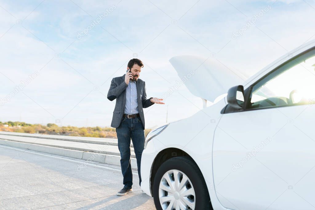 Adult man looking angry and talking on the phone at the side of the road. Latin man with his car hood open because of a mechanical problem