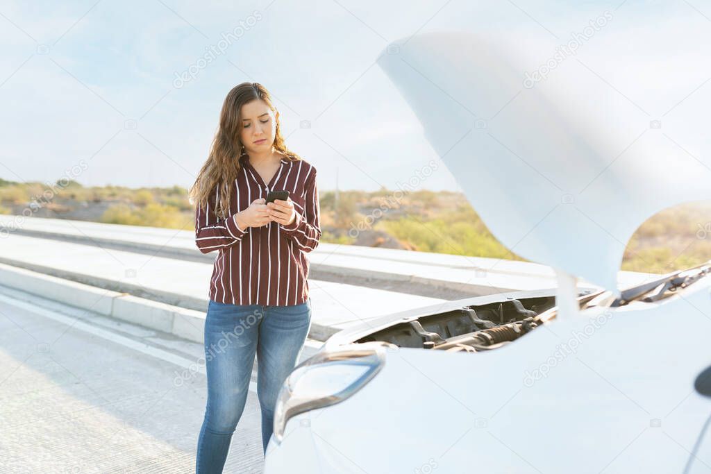 Sad young woman texting on her smartphone for road assistance help next to her broken-down car at the side of the highway