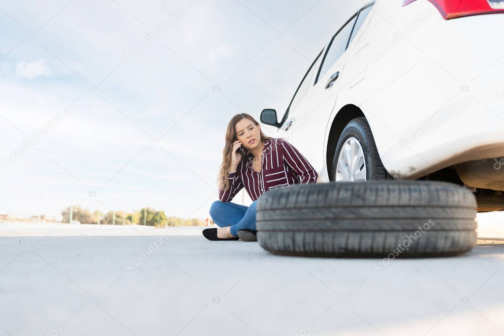 Pretty young woman with a flat tire sitting on the road next to her car. Woman calling for a tow truck and road assistance help